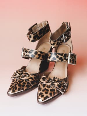 A pair of leopard print pointed-toe heels with triple buckle straps and silver metal toe caps, shot against a soft pink backdrop — equal parts vintage-inspired and unexpectedly wearable.