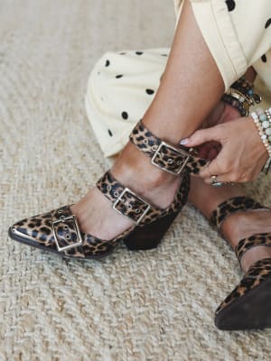 A close-up of leopard print block-heel booties with triple silver buckle straps being fastened by hand, styled with a cream polka dot skirt and stacked boho bracelets on a woven jute rug — the kind of shoe detail shot that makes you rethink everythin