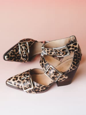 A pair of leopard print block heel booties with double silver buckle straps and a pointed toe, styled against a soft blush pink background — the kind of shoe that does all the talking.