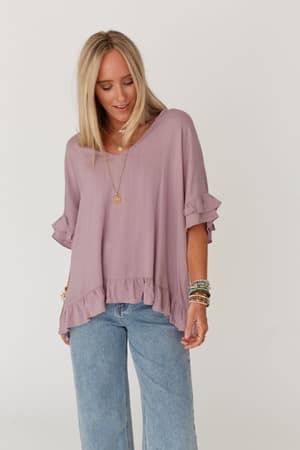 A blonde woman wears The Nest Let Me Be Ruffled Top in Pale Plum, a lightweight stretch knit blouse with ruffle details and a v-neckline, paired with light blue jeans. She stands against a white background, smiling subtly with layered necklaces and bracelets.