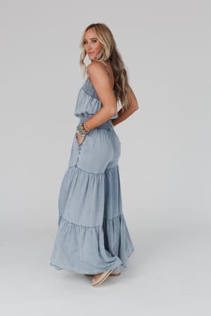 A woman with long blonde hair wears the Harlow Wide Leg Chambray Jumpsuit in blue, standing sideways and glancing over her shoulder. She accessorizes with bracelets, channeling a relaxed boho vibe against a plain white background.
