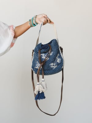 A hand holds up a chunky crochet bucket bag in denim blue with white floral embroidery, cream tassels, and a brown leather crossbody strap — accessorized with a stack of turquoise and gold beaded bracelets that feel just as considered as the bag itse