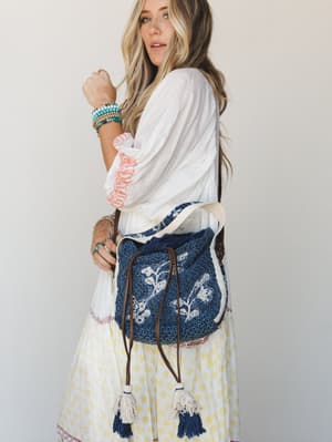 A sun-warmed model carries a navy crochet bucket bag with floral embroidery and tassel drawstring closures, styled with a breezy white embroidered blouse, a floral midi skirt, and a stack of turquoise bead bracelets — the whole look feels like a farm