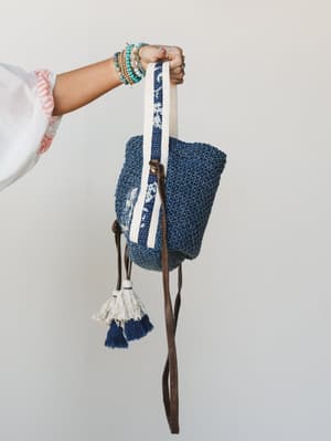 A hand holds up a chunky woven denim-blue crossbody bag with cream and navy tassel charms, a printed fabric strap, and a leather crossbody strap — layered bracelets on the wrist make it feel effortlessly personal.