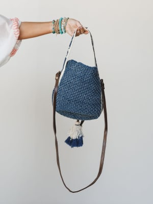 A hand stacked with turquoise and gold bracelets holds up a chunky crochet bucket bag in denim blue, finished with a leather crossbody strap and a pair of cream and navy tassels — the kind of bag that does all the talking.