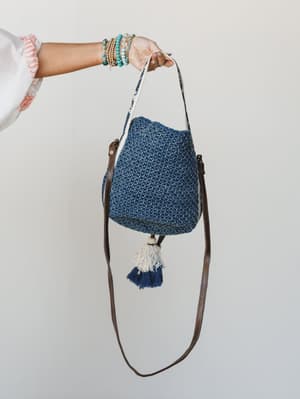 A hand stacked with turquoise and gold bracelets holds up a chunky crochet bucket bag in denim blue, finished with a leather crossbody strap and a pair of cream and navy tassels — the kind of bag that does all the talking.