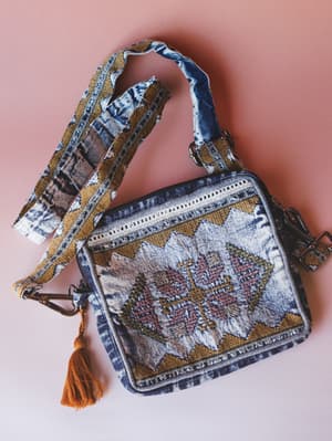 A structured denim crossbody bag laid flat against a dusty rose background, covered in tribal-style embroidery and geometric patchwork with a wide acid-wash strap and a warm amber tassel — the kind of bag that looks like it's been everywhere and want
