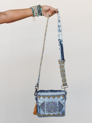 A beaded-bracelet-stacked hand holds up a denim crossbody bag with an acid-wash finish, tribal embroidery, and a tassel detail — the kind of bag that makes a casual outfit feel intentional.