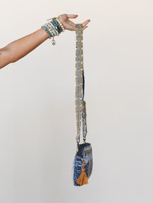 A stacked-bracelet wrist holds up a denim crossbody bag by its woven boho strap — the kind of bag that looks like it's been everywhere and has stories to tell.