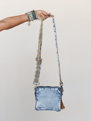 A hand stacked with beaded boho bracelets holds up a small acid-wash denim crossbody bag with a patterned guitar strap and tassel zipper pull — the whole thing feels like a summer road trip waiting to happen.