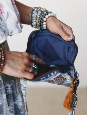 A close-up lifestyle shot of a denim patchwork crossbody bag held open by a heavily-stacked model with beaded bracelets and statement rings, showing off the navy interior lining and mustard tassel hardware — the kind of bag that looks like it's alrea