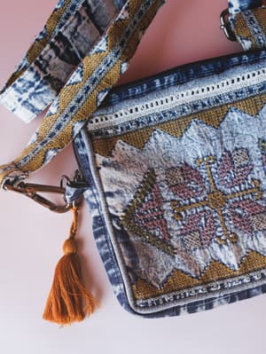 A close-up of a boho crossbody bag in acid-washed denim with geometric embroidery, mustard trim, lace zipper detail, and a burnt orange tassel charm — laid flat against a soft pink background.