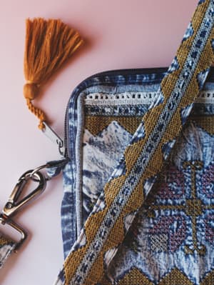 A close-up detail shot of a boho-style crossbody bag in washed indigo and cream, layered with a mustard embroidered woven strap and finished with a tassel zipper pull — the kind of bag that makes a whole outfit feel intentional.