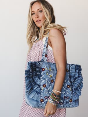 A sun-kissed blonde model holds an oversized embroidered denim tote with ruffle detailing against a clean light backdrop, styled with a pink geometric-print dress and stacked beaded bracelets — the kind of bag that makes a whole outfit feel more inte
