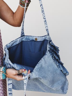 A roomy acid-wash denim tote held open to show its navy-lined interior — frayed ruffle trim along the sides, stacked beaded bracelets on both wrists, and a brass snap closure that makes the whole thing feel collected and well-loved.