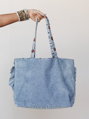 A washed denim tote bag with ruffle side panels and floral-embroidered straps, held up by a hand stacked with beaded bracelets against a soft neutral backdrop — the kind of bag that goes everywhere without trying.