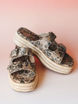 A pair of leopard print platform slide sandals with double buckle straps, silver grommet studding, and a woven raffia-wrapped platform sole, shot against a warm blush pink gradient background.
