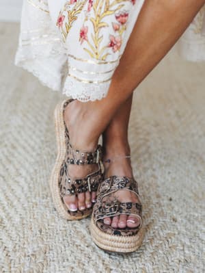 A close-up of leopard print platform slides with studded buckle straps and a woven espadrille sole, styled with a white floral-print wide-leg pant trimmed in lace and gold detail — the kind of shoe that does all the talking.