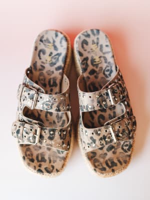 A pair of leopard-print platform slides shot overhead against a soft pink gradient background — double buckle straps with silver stud detailing and a jute-wrapped sole give them that lived-in, expressive energy.