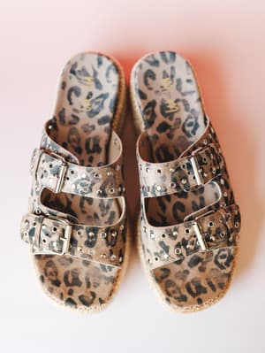 A pair of leopard-print platform slides shot overhead against a soft pink gradient background — double buckle straps with silver stud detailing and a jute-wrapped sole give them that lived-in, expressive energy.