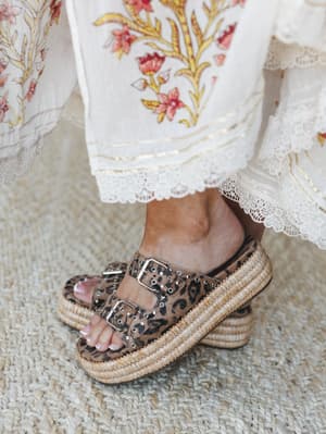 A close-up detail shot of leopard print platform sandals with woven raffia soles and silver buckle hardware, peeking out from beneath the lace-trimmed hem of a cream floral print skirt — the kind of styling moment that makes the whole outfit.