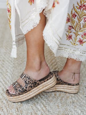 A close-up lifestyle shot focused on leopard print platform slides with woven raffia soles, styled against the lace-trimmed hem of a cream floral maxi dress — the kind of pairing that looks effortless but definitely wasn't an accident.