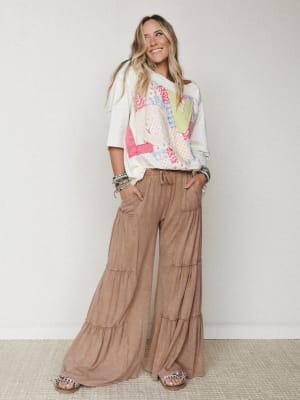 A relaxed full-body shot of a smiling model in flowy tiered wide-leg pants and an oversized patchwork graphic tee, layered with stacked bracelets — the kind of effortless outfit that looks like it took no effort but absolutely did.