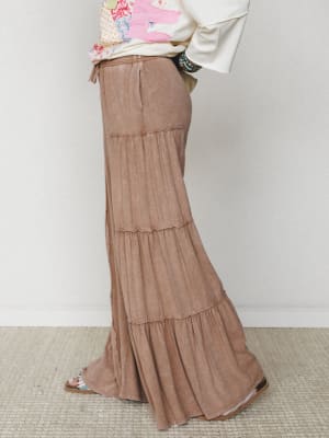 A side-profile shot of a flowy, tiered wide-leg pant in washed mocha, styled with a cream patchwork top and stacked turquoise bracelets — the kind of outfit that looks effortless but absolutely wasn't an accident.