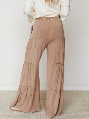 Shot from behind, a model wears dramatically tiered wide-leg pants in a washed mocha tone paired with a cream long-sleeve top — relaxed, earthy, and full of easy movement.