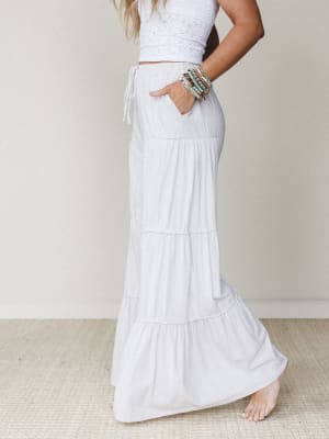 A sun-kissed model poses in profile wearing flowy white tiered wide-leg pants and a cropped crochet top, stacked beaded bracelets catching the light — the whole look feels like a slow morning somewhere warm.
