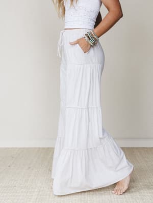 A sun-kissed model poses in profile wearing flowy white tiered wide-leg pants and a cropped crochet top, stacked beaded bracelets catching the light — the whole look feels like a slow morning somewhere warm.