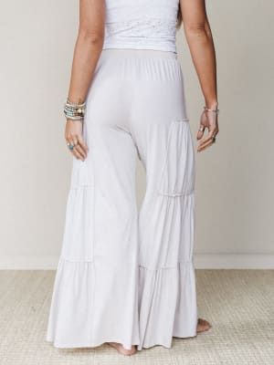 Shot from the back, a model wears flowy tiered wide-leg pants in a soft natural white paired with a white eyelet crop top — the kind of effortless, breezy outfit that feels equally at home on a beach boardwalk or a summer patio.