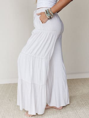A side-profile shot of flowy tiered wide-leg pants in a soft natural white, styled with a cropped eyelet top and stacked beaded bracelets — the kind of outfit that looks equally at home on a beach boardwalk or a slow Sunday morning.
