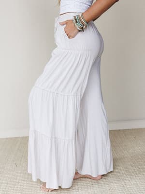 A side-profile shot of flowy tiered wide-leg pants in a soft natural white, styled with a cropped eyelet top and stacked beaded bracelets — the kind of outfit that looks equally at home on a beach boardwalk or a slow Sunday morning.