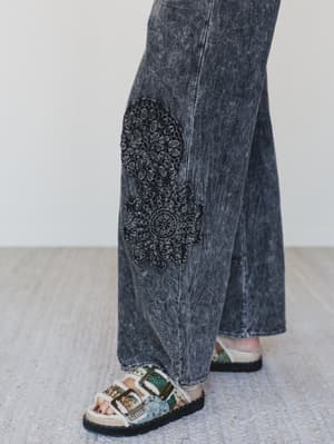 Person wearing acid-wash jeans with a decorative patch and patterned sandals on a light wooden floor.