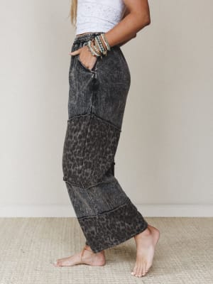 A side-profile shot of wide-leg pants in an acid-washed ash black leopard print, styled with a white crochet crop top and stacked beaded bracelets — relaxed and expressive with a lived-in edge.