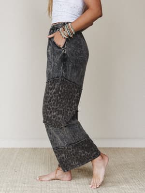 A side-profile shot of wide-leg pants in an acid-washed ash black leopard print, styled with a white crochet crop top and stacked beaded bracelets — relaxed and expressive with a lived-in edge.