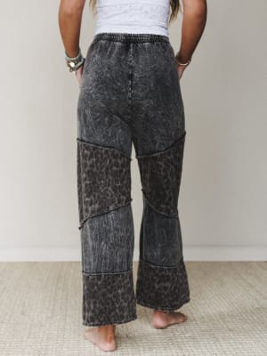 Shot from behind, these wide-leg pants in ash black mineral wash feature alternating patchwork panels of acid wash and leopard print, styled with a white eyelet top and stacked turquoise bracelets for a lived-in boho vibe.