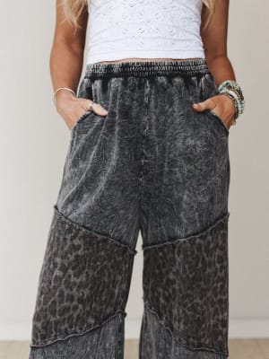 A close-up mid-shot of wide-leg pants in an acid-washed ash black with a subtle leopard print that reveals itself in the lower panels — styled with a white eyelet crop top and stacked beaded bracelets for a relaxed, expressive vibe.