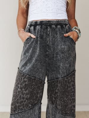 A close-up mid-shot of wide-leg pants in an acid-washed ash black with a subtle leopard print that reveals itself in the lower panels — styled with a white eyelet crop top and stacked beaded bracelets for a relaxed, expressive vibe.