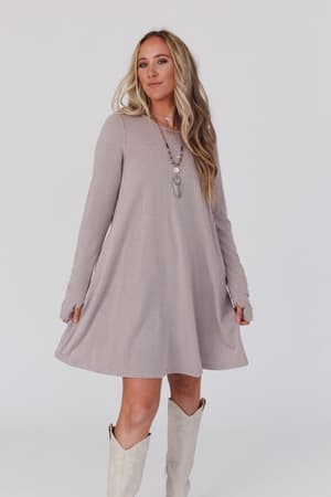 A woman with long blonde hair poses against a white background wearing The Nest Jake Long Sleeve Crewneck Dress in Mocha Latte, styled with white knee-high boots and layered necklaces.