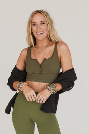 A woman with long blonde hair wears the Hook It Up Ribbed Brami in olive, paired with matching leggings and accessorized with layered necklaces, bracelets, and rings. She smiles, draping a black shirt loosely over her arms.