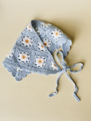 A sky blue crochet granny square headband bandana laid flat, detailed with white daisy motifs and scalloped edges, finishing in long tie strings — the kind of thing you'd spot at a farmers market and immediately ask where she got it.