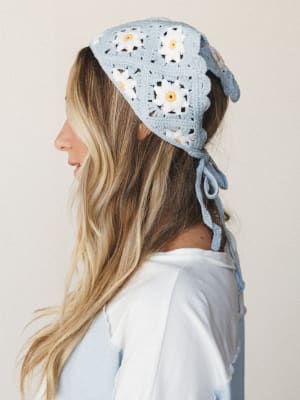 A side-profile close-up of a crochet granny square headband in soft sky blue with white daisy motifs and yellow centers, tied at the back with trailing yarn strings — sweet, handmade-feeling, and completely the kind of thing you'd wear to a farmers m