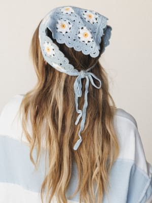 Shot from behind, a model wears a soft sky blue crochet headscarf with granny square daisy motifs and a trailing tie closure — the kind of piece that makes a ponytail feel intentional.