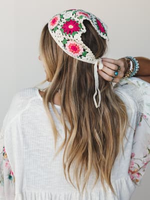 Shot from behind, a model ties a crochet granny square headband into her wavy blonde hair — the kind of accessory that makes a white gauze top feel like a whole personality.