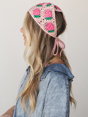 A sun-warmed side profile shot of a model wearing a crochet granny square headscarf covered in strawberry motifs — tied at the nape and layered over a denim shirt, the whole thing feels like summer at a farm stand.
