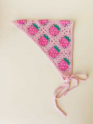 A crochet triangle bandana headband in soft pink, covered in granny-square strawberry motifs with bright pink fruit, green leaves, and yellow seed details — laid flat on a cream background with tie strings trailing loose.