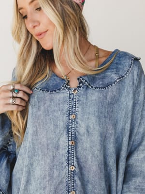 A close-up shot of a relaxed chambray button-front top with a peter pan collar and embroidered placket detail, styled with layered silver and turquoise rings, a delicate gold necklace, and a pink crochet floral headband — the whole thing has that eff