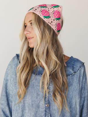 A crochet strawberry headband in blush pink sits on a model with wavy blonde hair, paired with a soft chambray button-down — the kind of accessory that makes a whole outfit feel intentional without trying too hard.